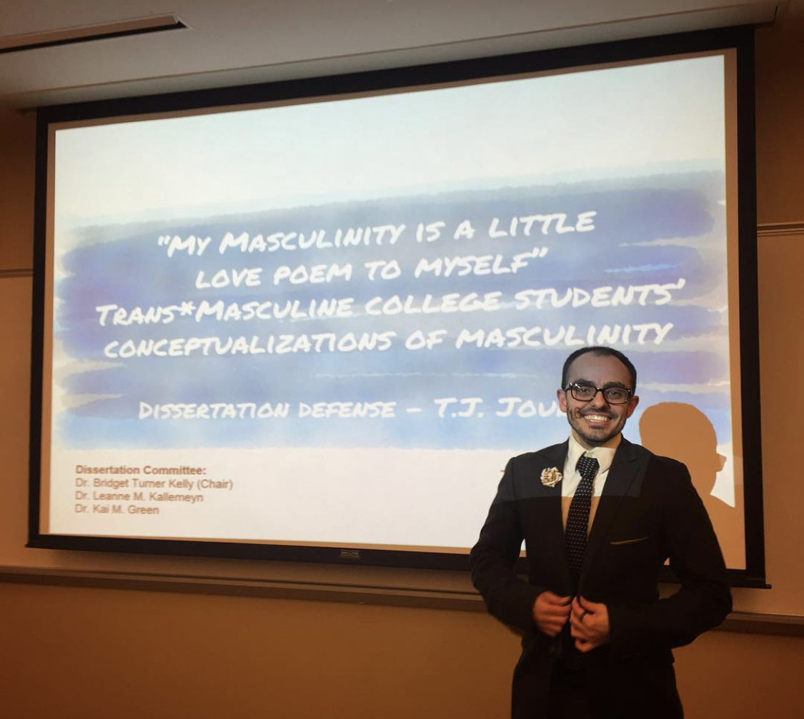 T.J. beaming with a smile and a brown suit in front of a large screen. The screen displays a slide with the title: "My Masculinity is a  Little Love Poem to Myself": Trans*Masculine College Students' Conceptualizations of Masculinity / Dissertation Defense - T.J. Jourian